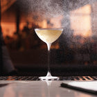 A clear stemmed cocktail coupe glass filled with a Silvertear vintage vodka martini set on a bar counter with a blurred warm-toned bar background and visible spray mist around the glass.