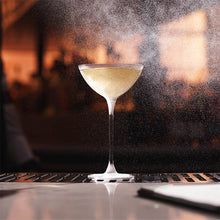 A clear stemmed cocktail coupe glass filled with a Silvertear vintage vodka martini set on a bar counter with a blurred warm-toned bar background and visible spray mist around the glass.