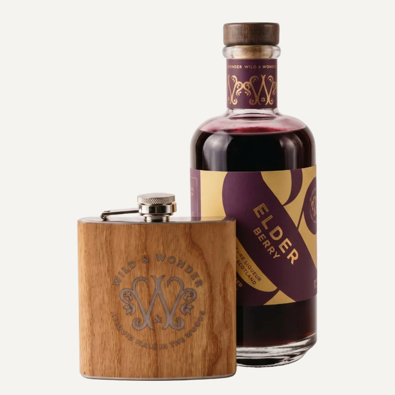 A bottle of Elder Berry by Wild & Wonder next to a matching wooden flask with an engraved Wild & Wonder monogram, set against a clean off-white background.