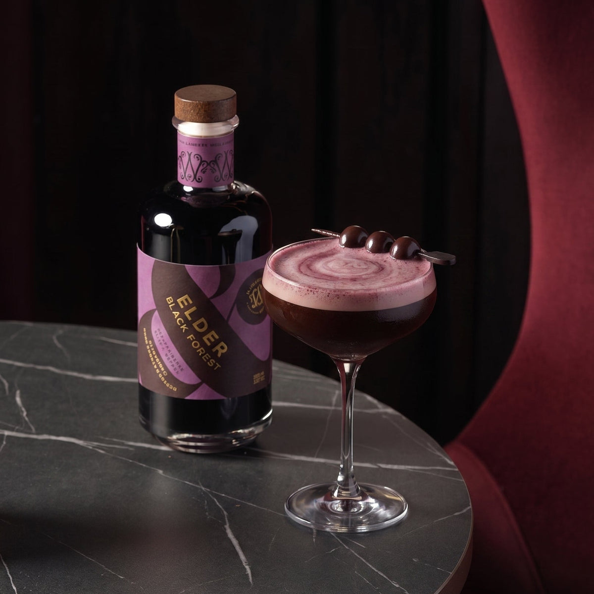 A bottle of Elder Black Forest liqueur stands beside a coupe glass filled with a dark berry espresso martini topped with pink foam and garnished with cherries, set on a dark marble table against a moody black and burgundy background.