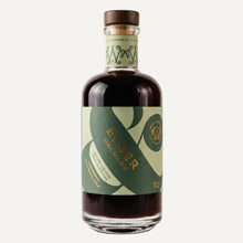 A bottle of Elder Orchard liqueur with a dark amber liquid, featuring a green and light green label with gold lettering and a brown wooden cap, set against a plain light grey background.