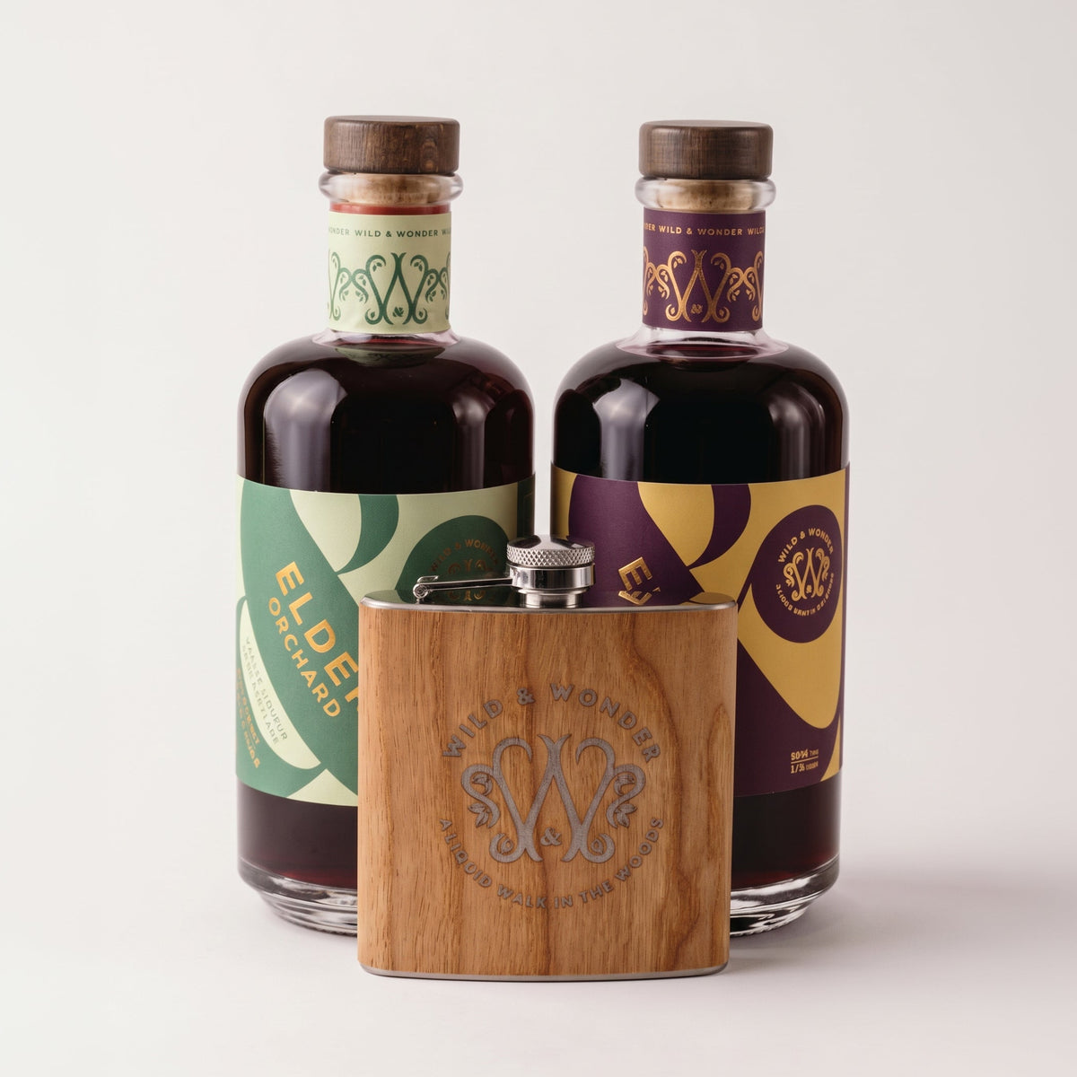 A bottle of Elder Orchard and Elder Berry liqueur with decorative labels and a small wooden hip flask in front, photographed on a clean light grey studio background. The labels show the brand name Wild & Wonder