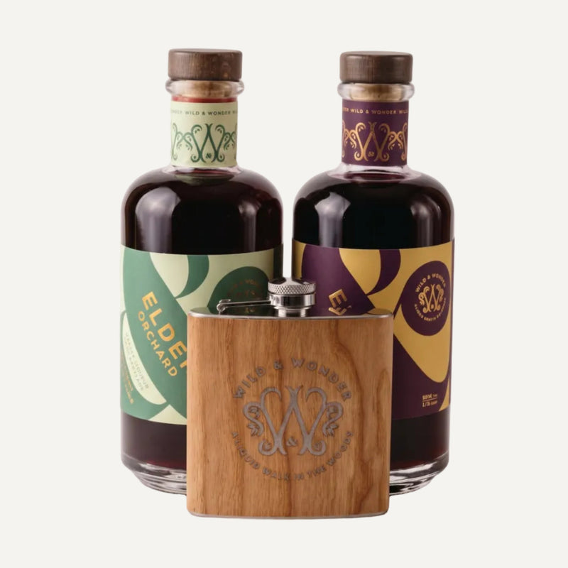 A bottle of Elder Orchard and Elder Berry liqueur with decorative labels and a small wooden hip flask in front, photographed on a clean light grey studio background. The labels show the brand name Wild & Wonder