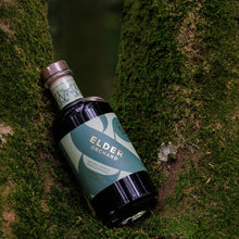 A bottle of Wild & Wonder Elder Orchard apple and bramble liqueur resting diagonally on mossy tree bark in an outdoor forest setting.