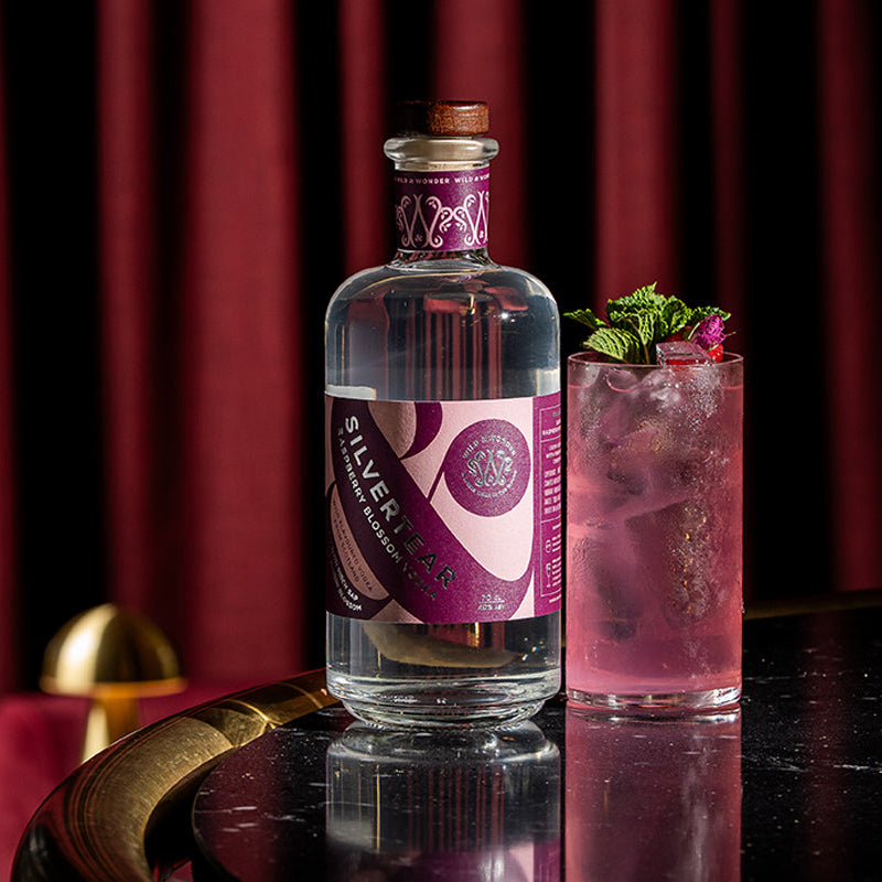 A bottle of Silvertear Raspberry Blossom vodka next to a pink cocktail in a highball glass, with deep red curtains behind