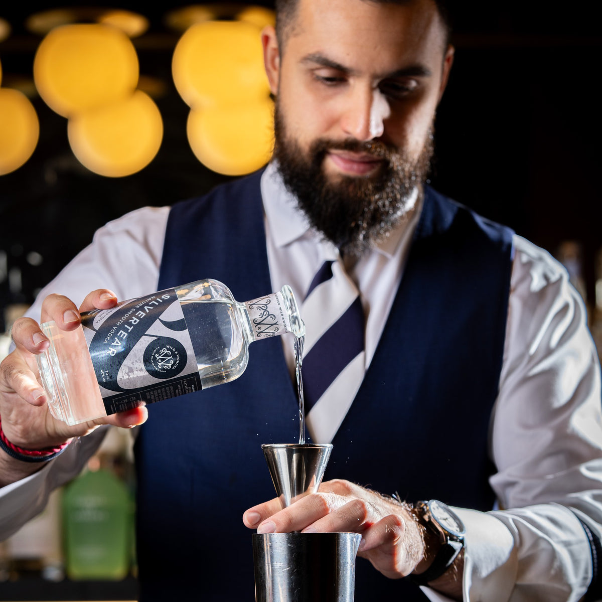 A bearded bartender in a navy waistcoat pours Silvertear Nature vodka into a jigger over a cocktail shaker, with warm globe lights behind.