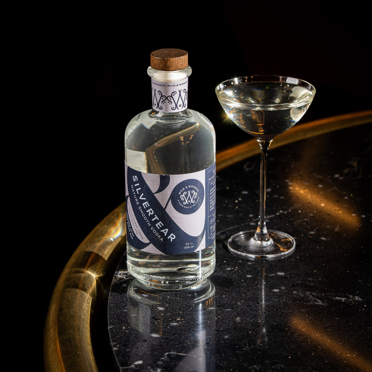 A bottle of Silvertear Nature smooth birch sap vodka next to a martini in a coupe glass on a curved black marble bar with a brass edge.