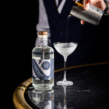 A bartender pours a Silvertear Nature vodka martini into a frosted coupe glass, with the bottle beside it on a black marble bar with a brass rail.