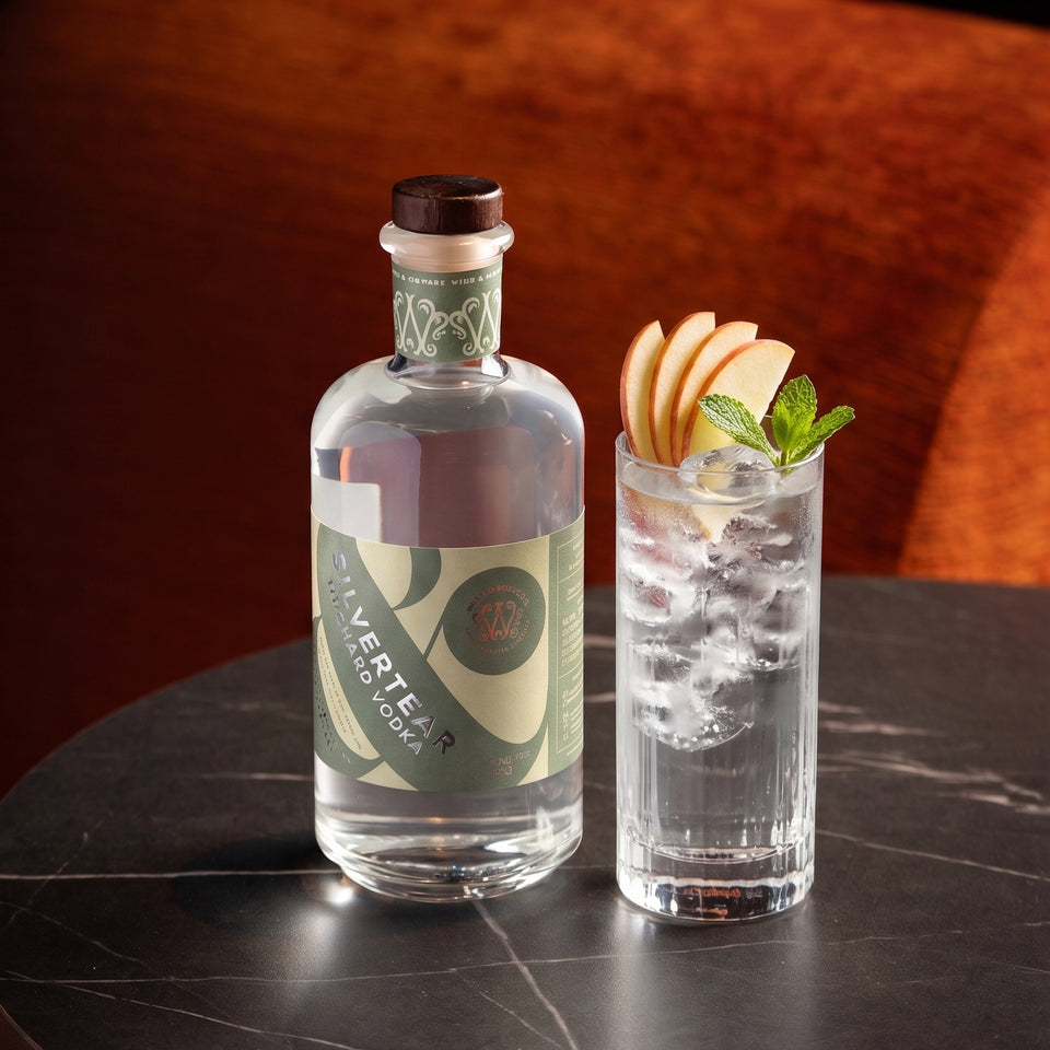 A bottle of Silvertear Orchard vodka next to a highball glass filled with a vodka and tonic, ice, apple slices, and mint, set on a dark marble table against a warm brown background.