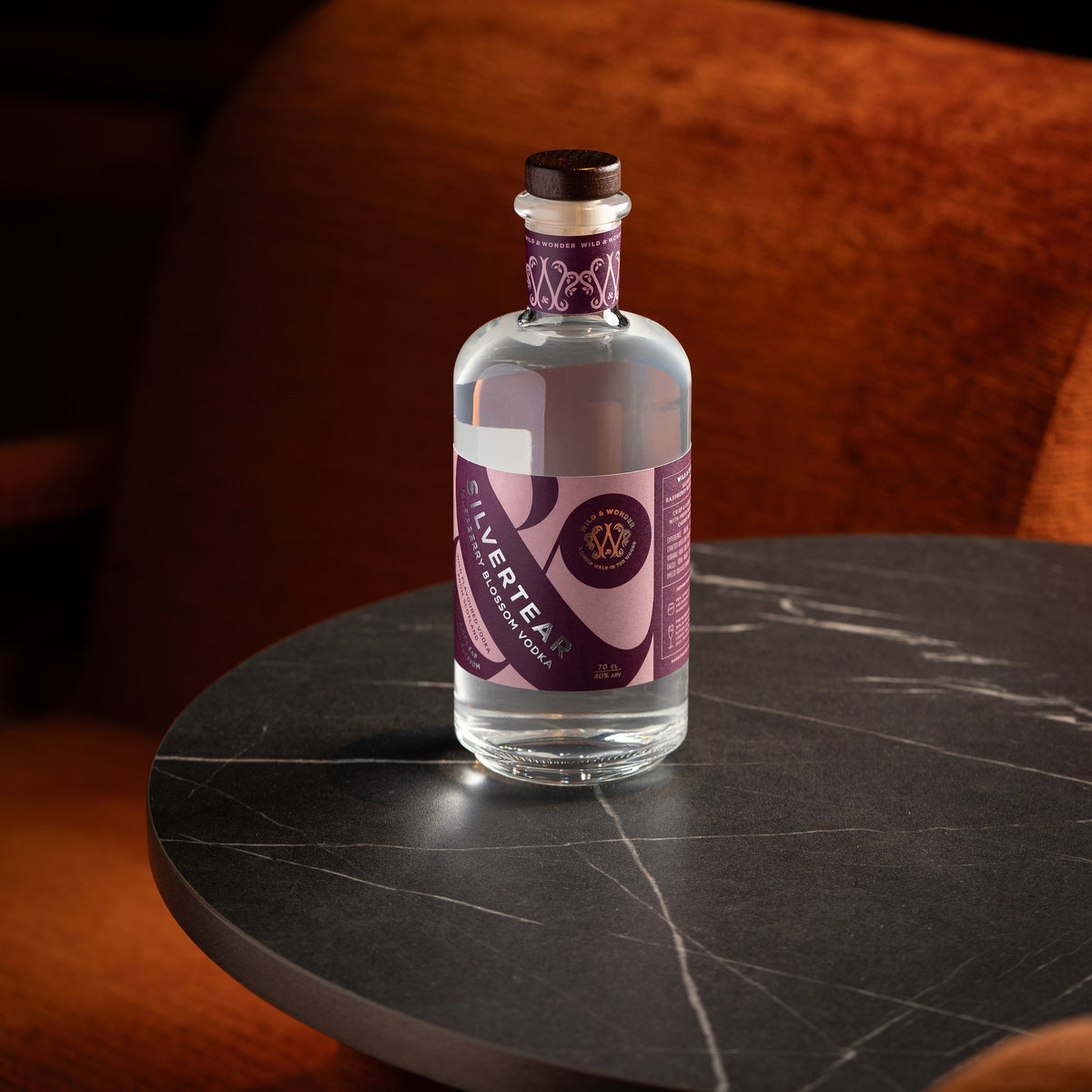 A bottle of Silvertear Raspberry Blossom vodka with its pink and purple label on a round black marble bar table, with a rust-orange velvet armchair behind it in a dimly lit bar.
