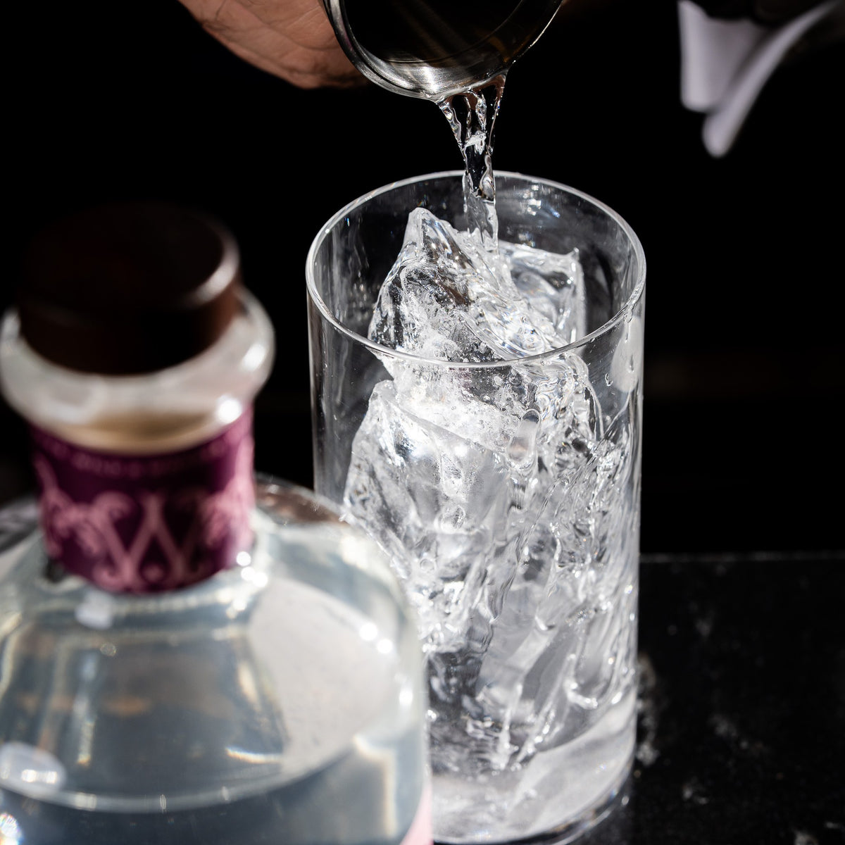 A bartender pours Silvertear Raspberry Blossom vodka over ice in a mixing glass, with the bottle in the foreground on a black marble bar.