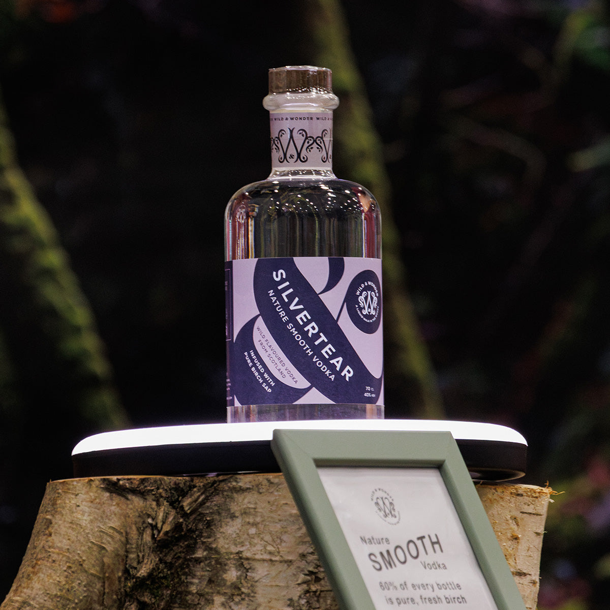 A bottle of Silvertear Nature smooth vodka displayed on rustic birch plinth with ring light, with a framed label in the foreground and a dark, blurred natural background.