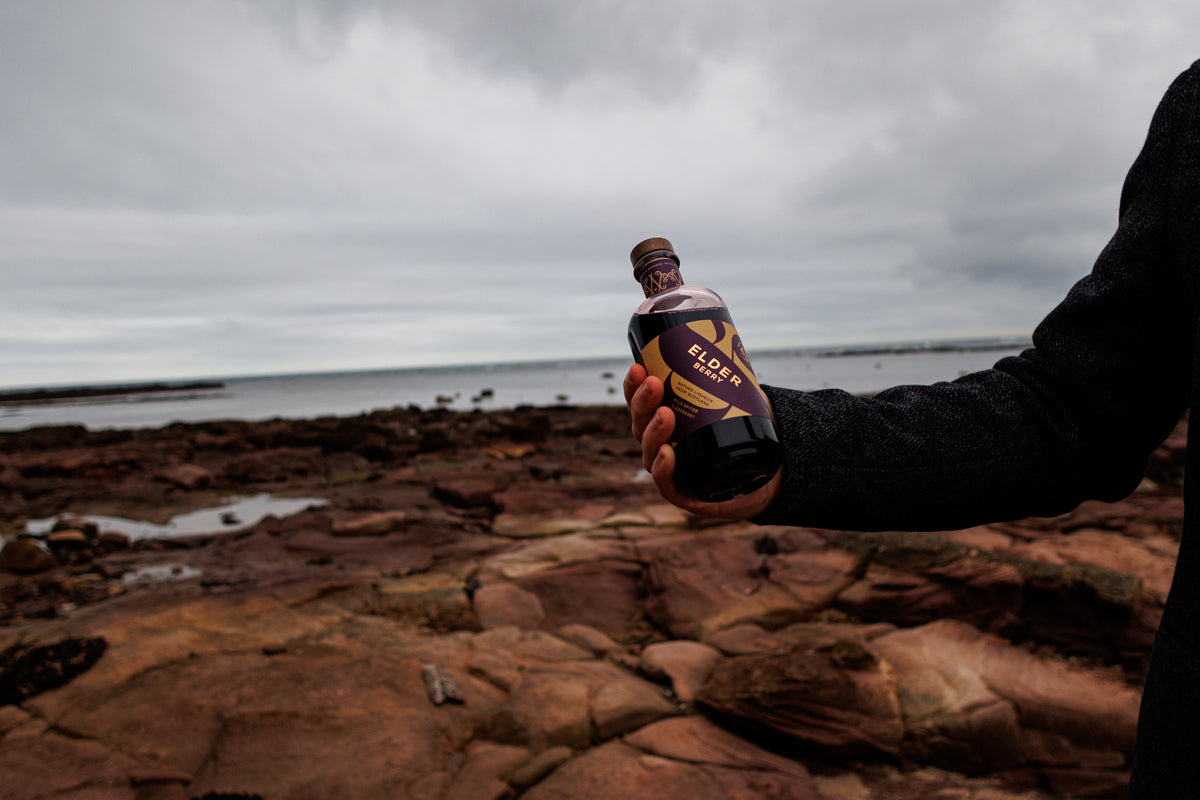 A hand holding up a bottle of Elder Berry liqueur on a rocky shore under a grey sky