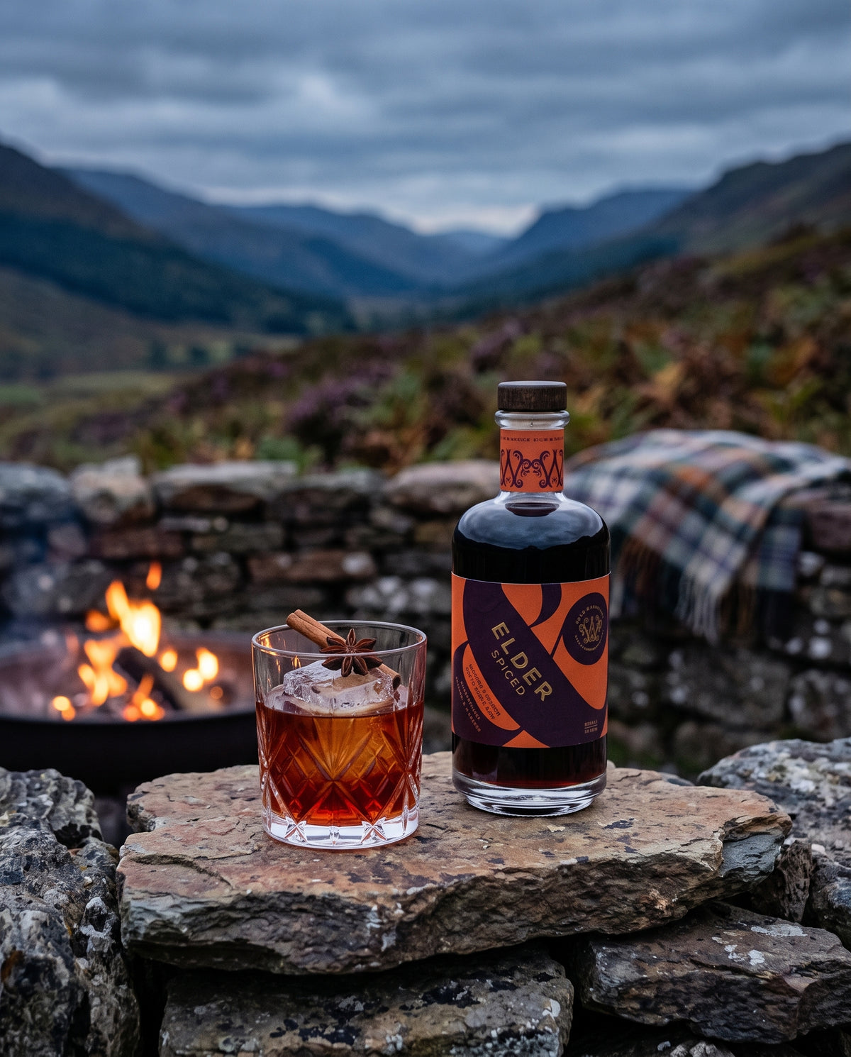 A bottle of Elder Spiced liqueur and a short drink with a cinnamon stick on a stone wall by a fire, looking out over a Highland glen
