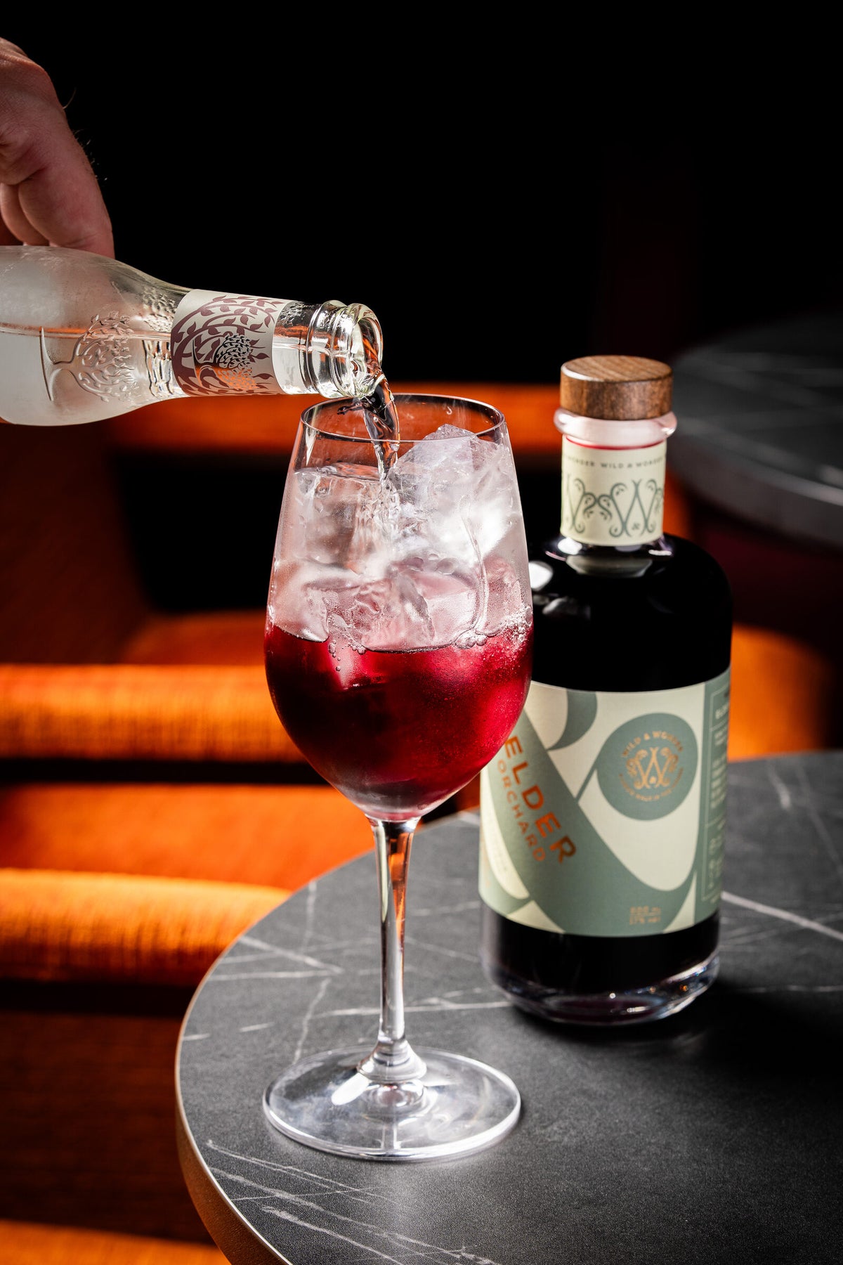 Tonic being poured into a dark berry spritz beside a bottle of Elder Orchard