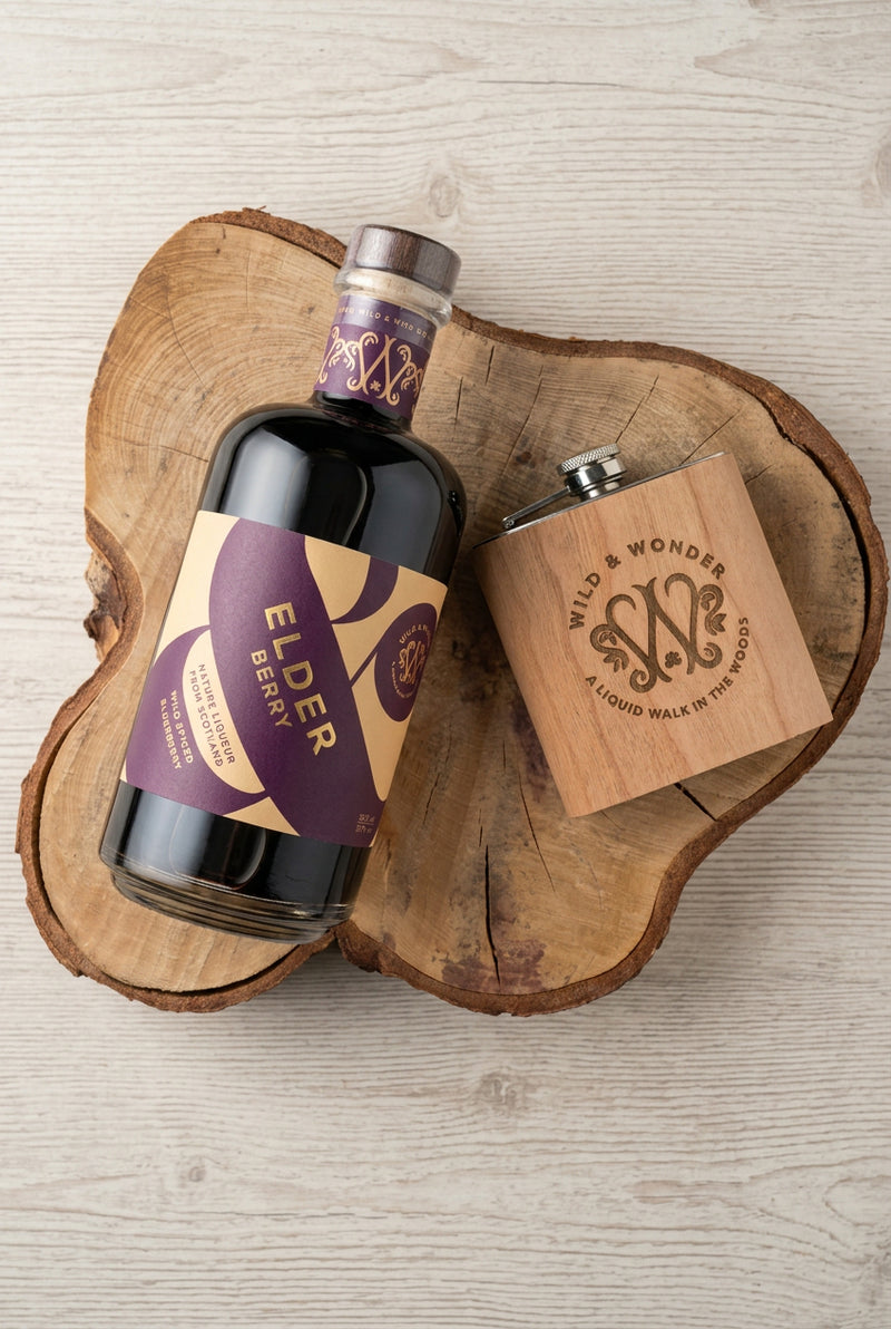 Bottle of Elder Berry liqueur and a wood-wrapped Wild & Wonder hipflask on a slice of tree trunk, seen from above