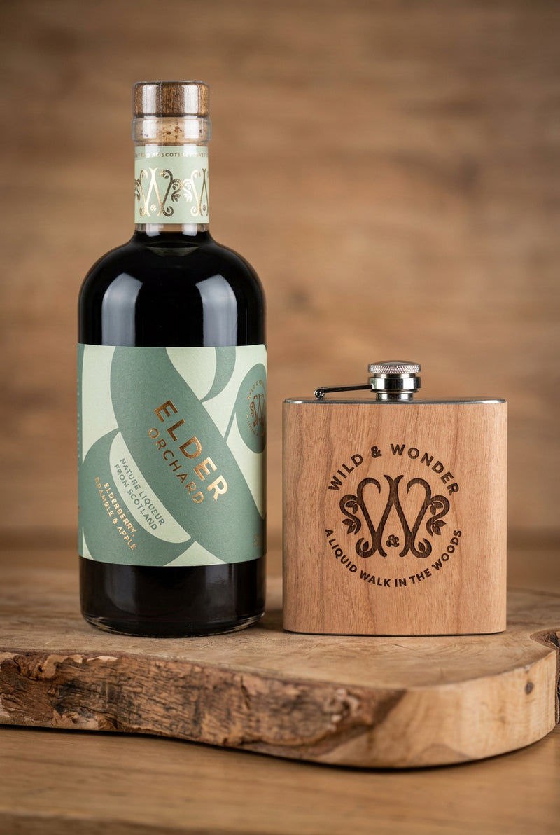 Bottle of Elder Orchard liqueur beside a wood-wrapped Wild & Wonder hipflask on a wooden board