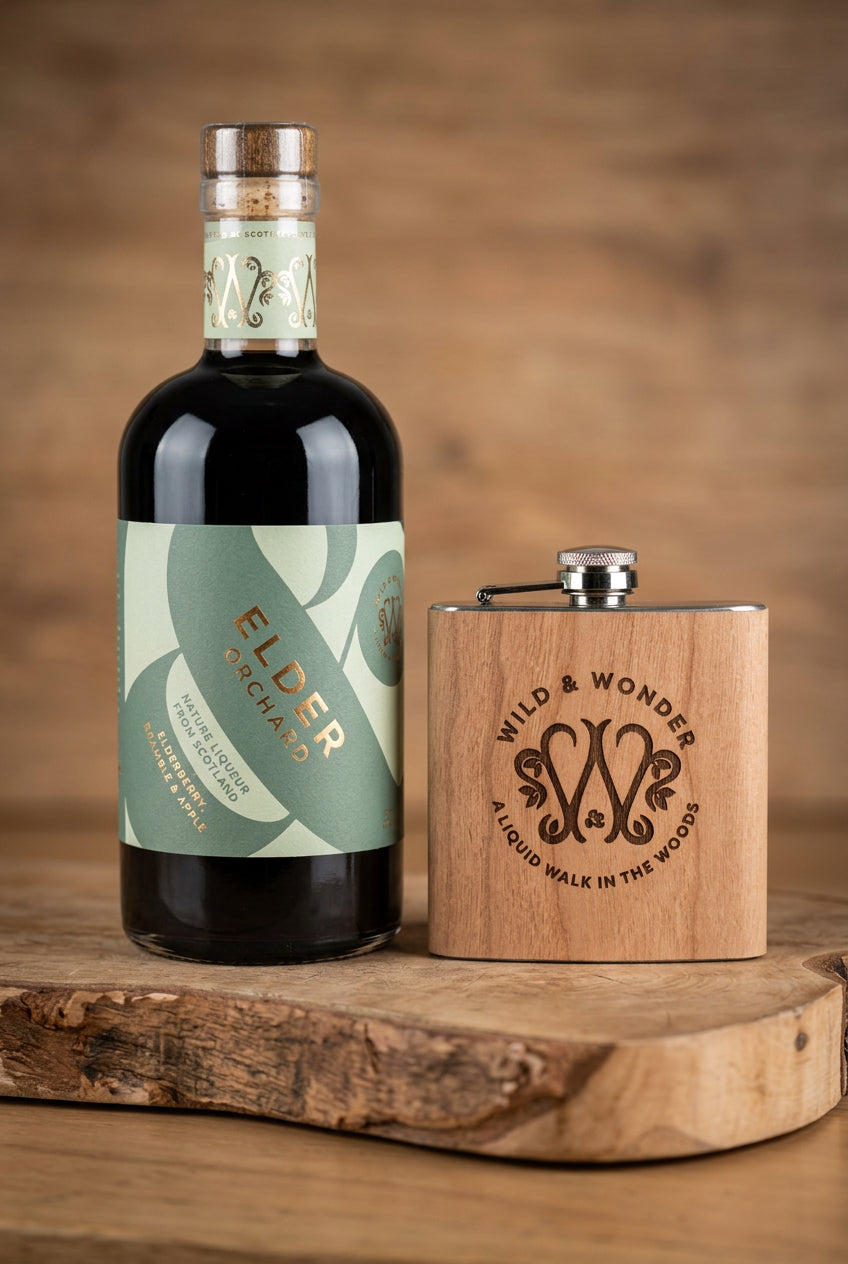 Bottle of Elder Orchard liqueur beside a wood-wrapped Wild & Wonder hipflask on a wooden board