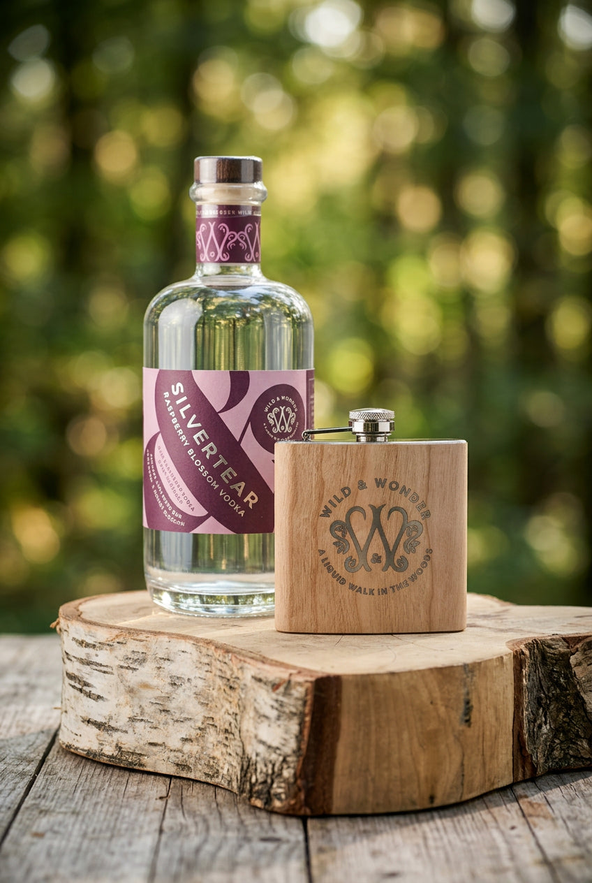 Silvertear Raspberry Blossom vodka beside a wood-wrapped Wild & Wonder hipflask on a birch log in a sunlit forest
